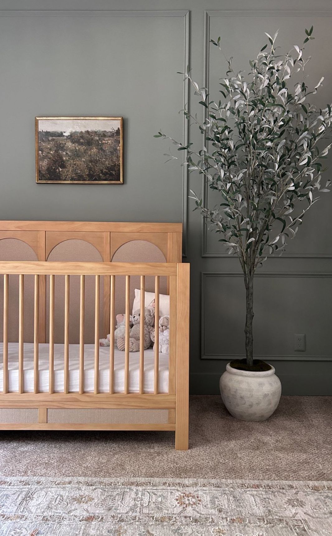 A boy nursery with olive green walls, a wooden crib, a faux tree, and a vintage landscape art print