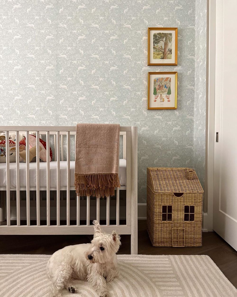 A nursery with dark wood floors, white and sage rabbit printed wallpaper, a white crib with a throw blanket and stuffed animals, and vintage art prints