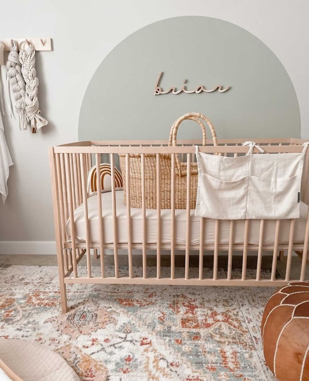 A boho themed nursery room with a sage wall accent, light wood crib, a boho rug, and other boho accents