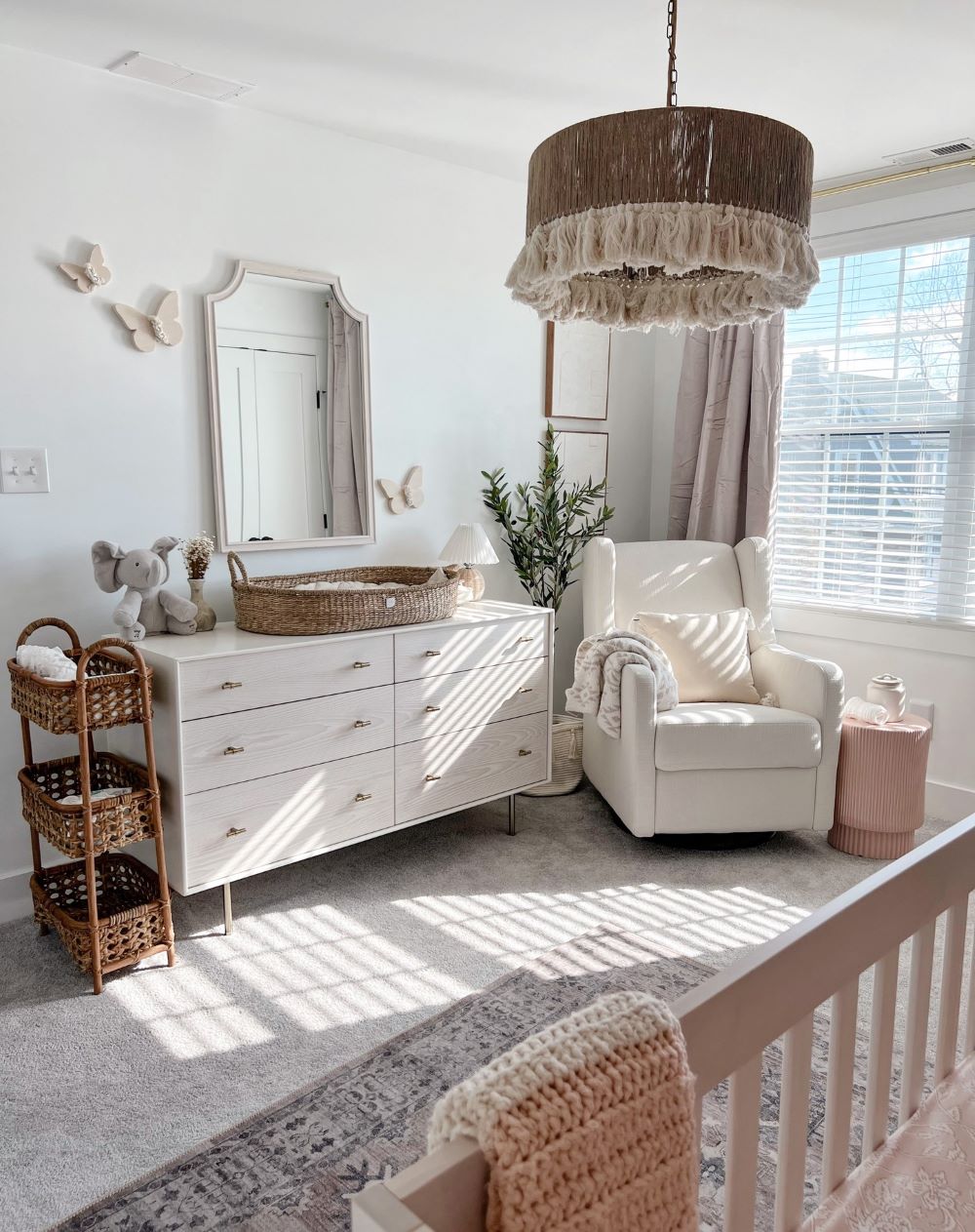 A girl's nursery room with white walls, a boho rug and other boho decor pieces, pink accents, and butterfly wall decor