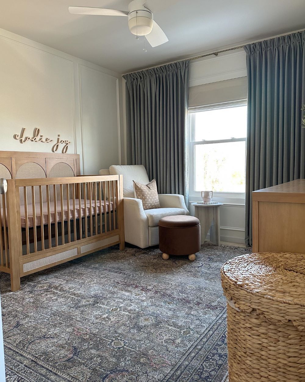 A girls nursery room with a dark blue boho style rug, light wooden furniture, white walls, and slate blue curtains