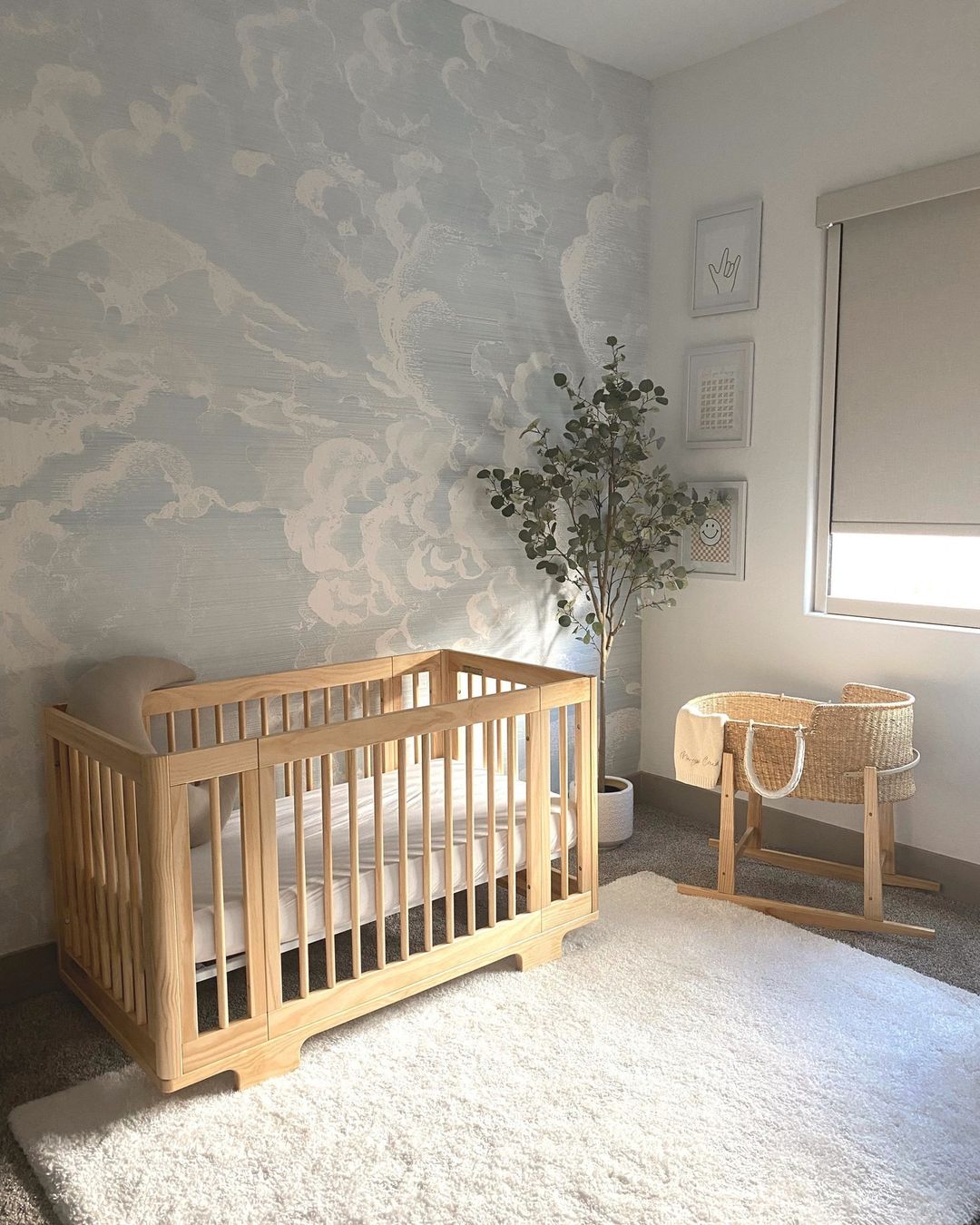 A boys nursery room with cloudy blue sky wallpaper, a faux plant, and light wood furniture