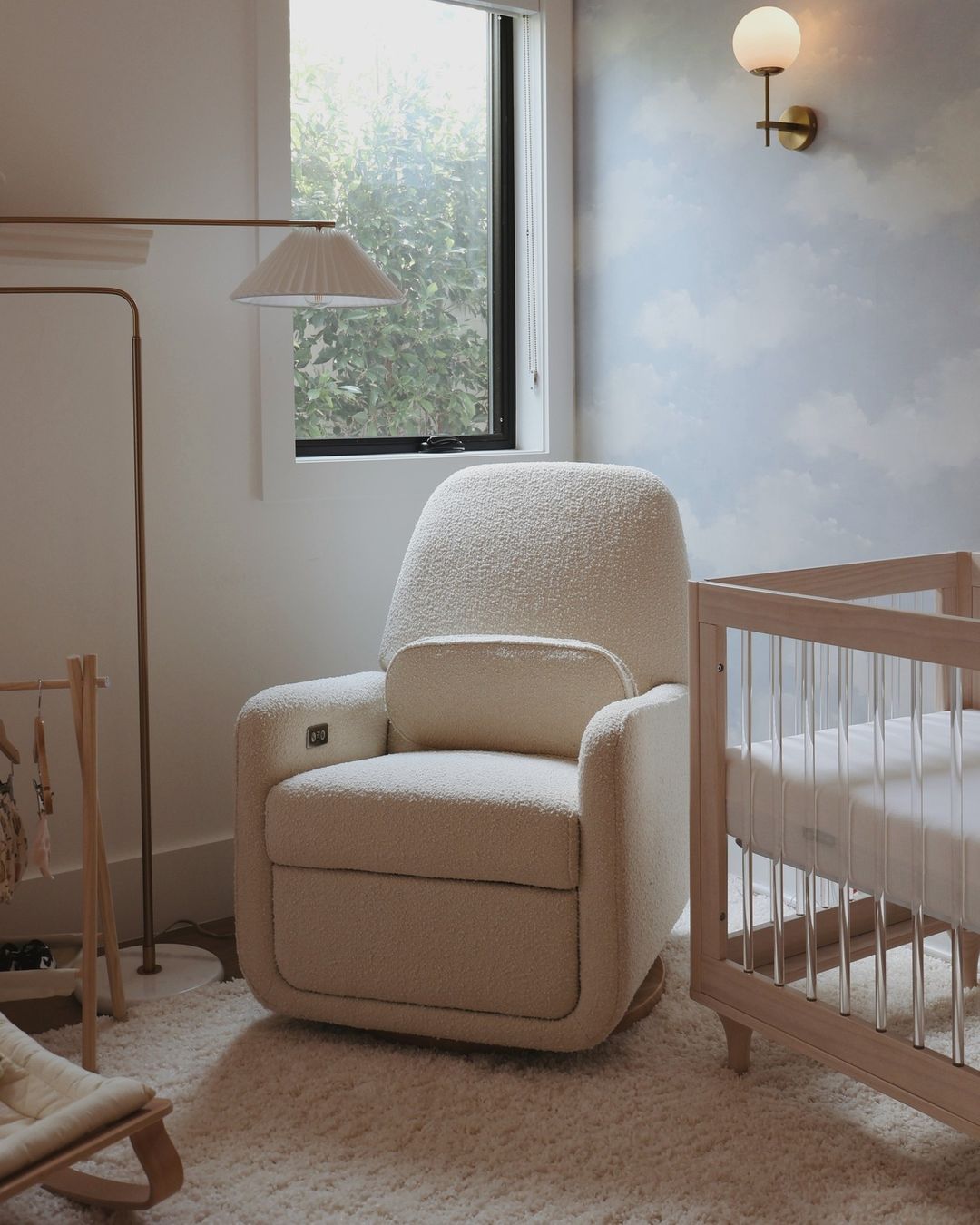 A baby boy nursery with cloudy blue sky wallpaper, a boucle glider, and low lighting