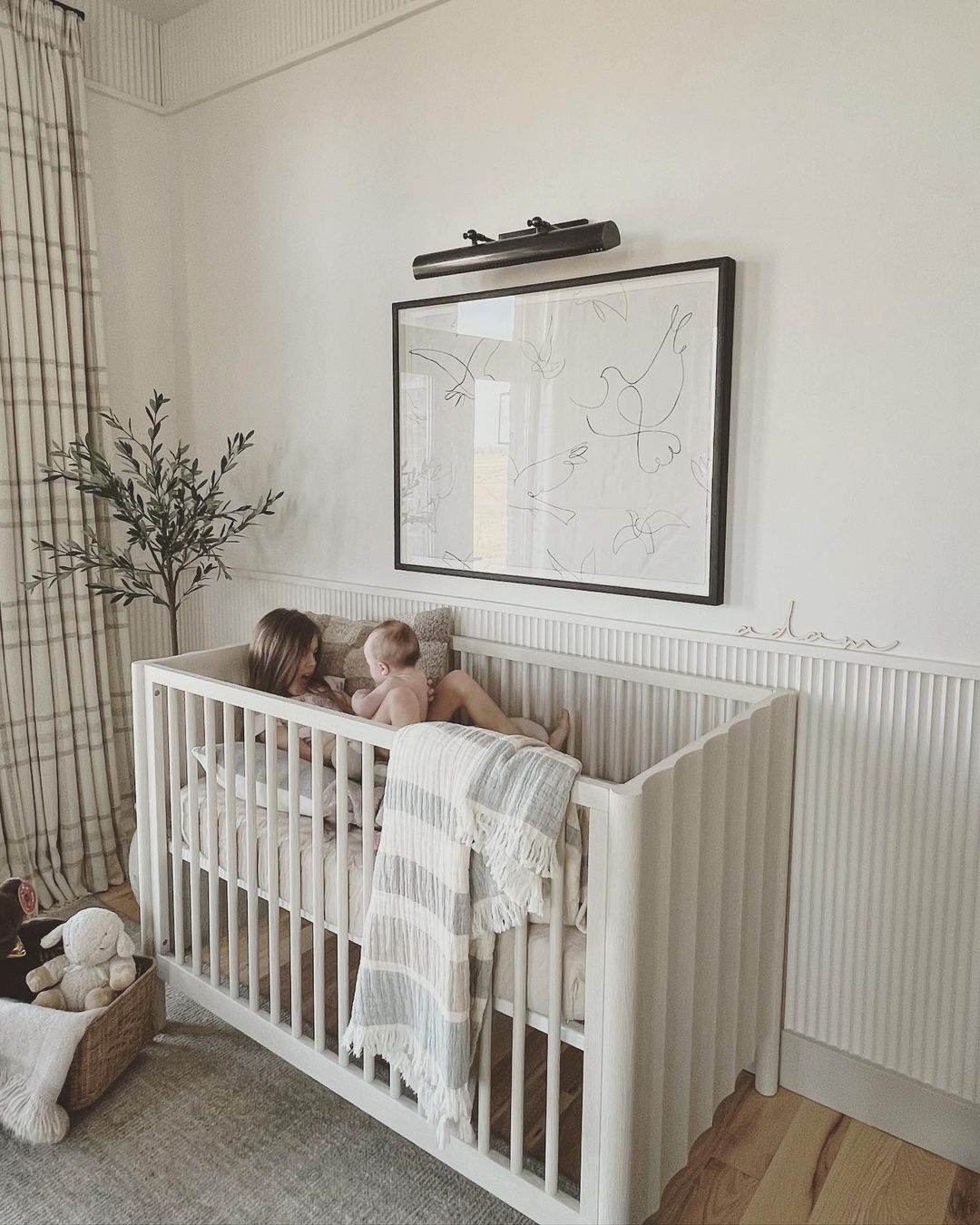 A baby boy nursery room with white walls and furniture, featuring a board and batten half wall, plaid curtains, and a faux plant