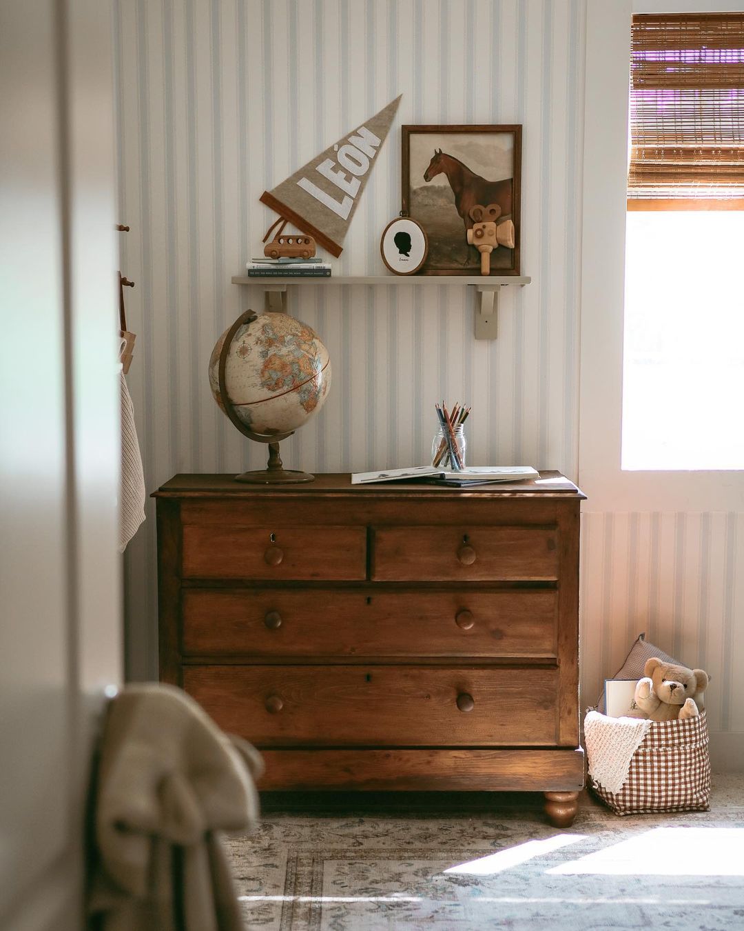 A baby boy nursery room with striped wallpaper, warm toned furniture, and decor details such as a globe, horse wall art, and more