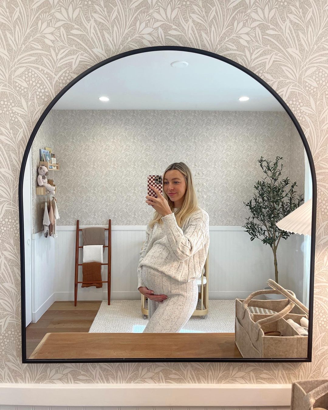 A pregnant women standing in front of the mirror of her daughters nursery featuring neutral floral wallpaper, white board and batten, and natural elements