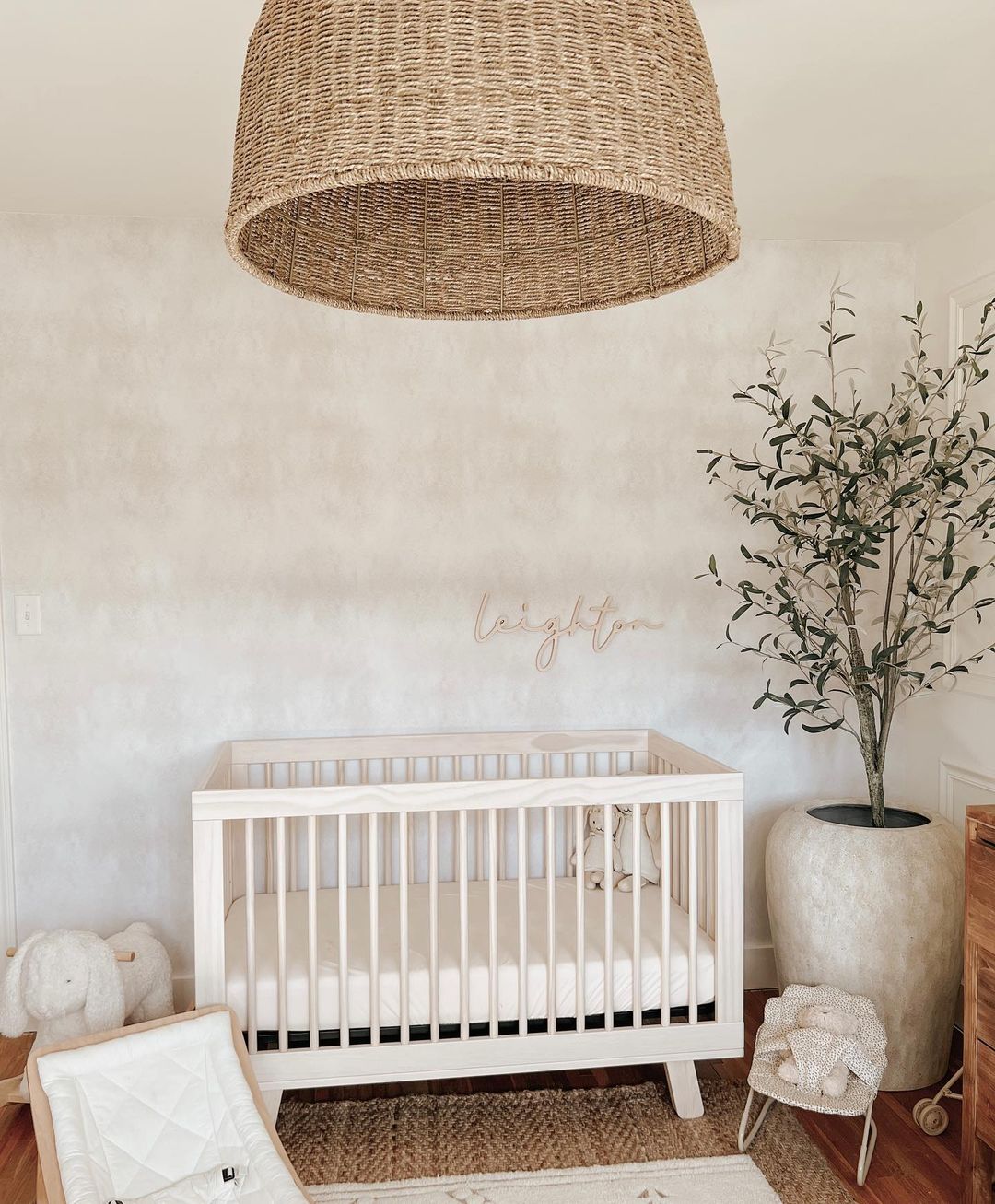 A girl's nursery featuring a brushed creamy beige wallpaper, plant decor, stuffed animals, and a raffia light fixture