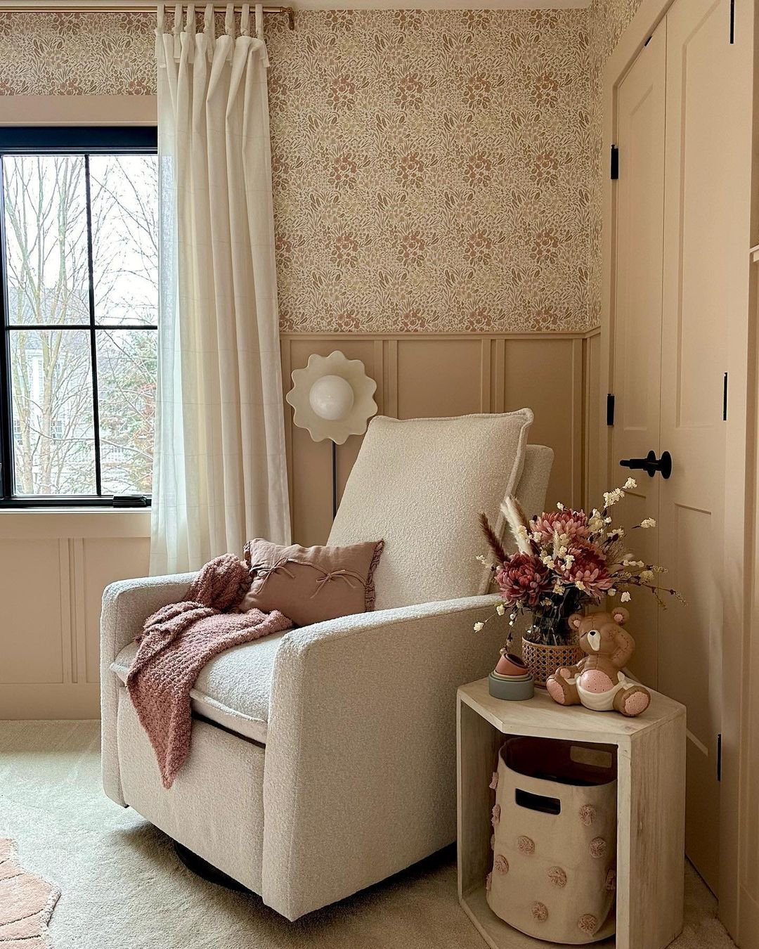 A girl's nursery with floral beige wallpaper and beige board and batten with white furniture and curtains and pops of pink and floral accents