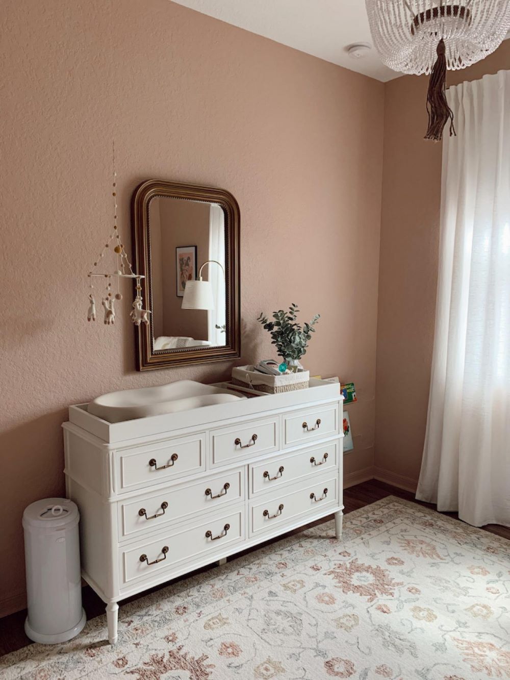 A nursery with soft pink walls, a white dresser, and boho decor pieces