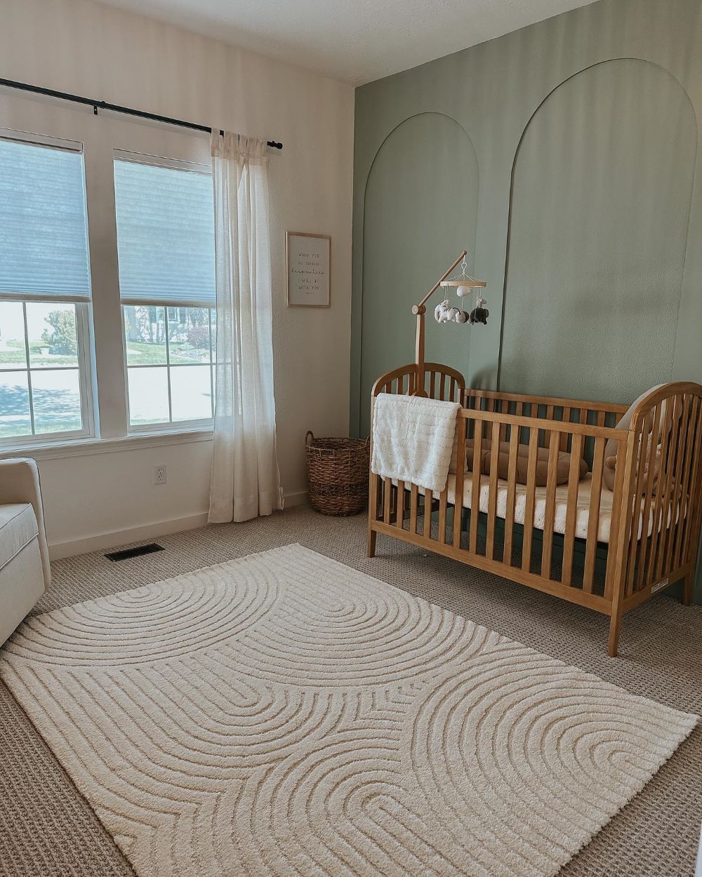 A nursery with soft green walls, a light wood crib, and boho decor pieces