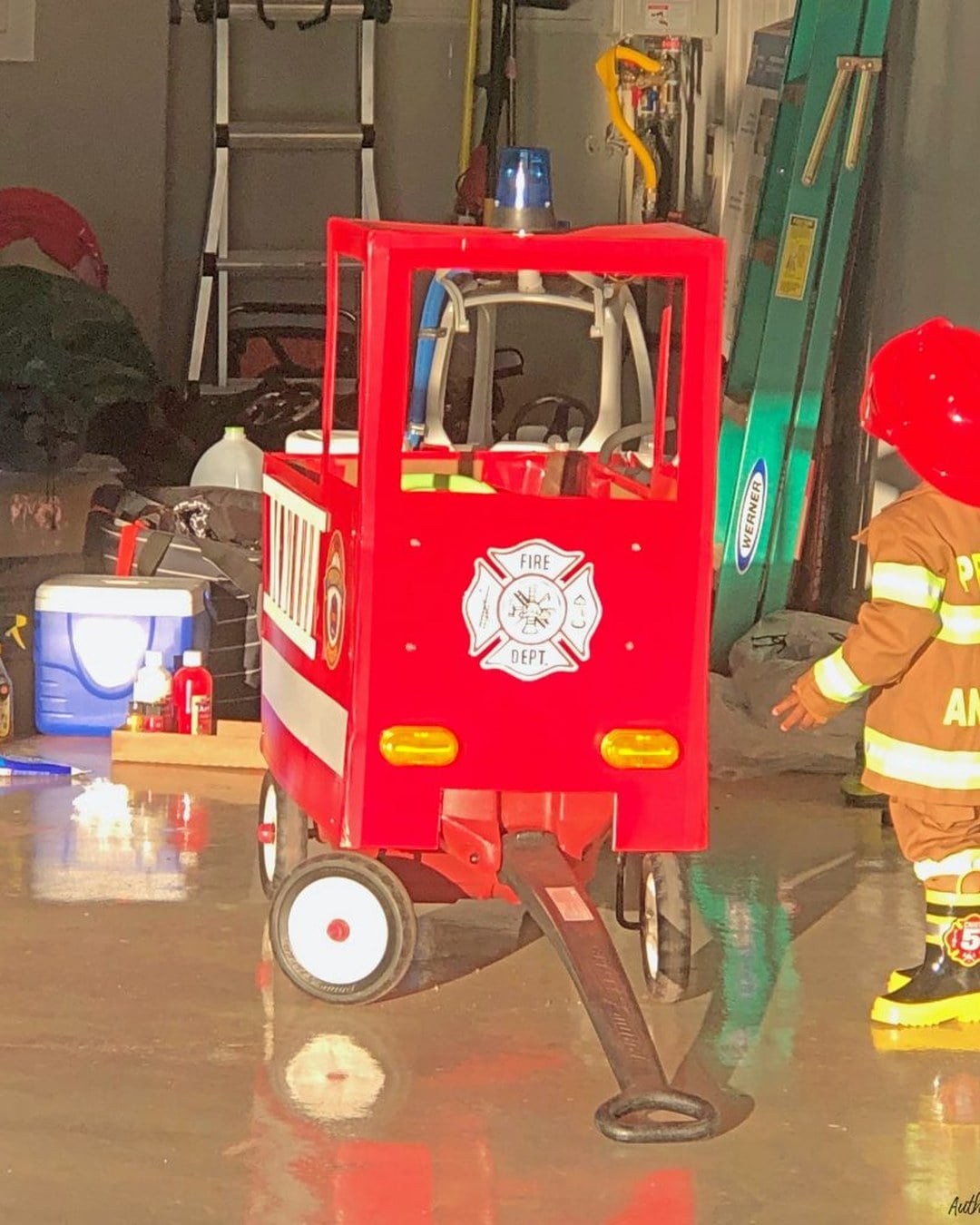 A toddler wearing a handmade red firefighter coat and helmet next to a baby in a white-and-black spotted dalmatian onesie, with a cardboard fire truck prop nearby.