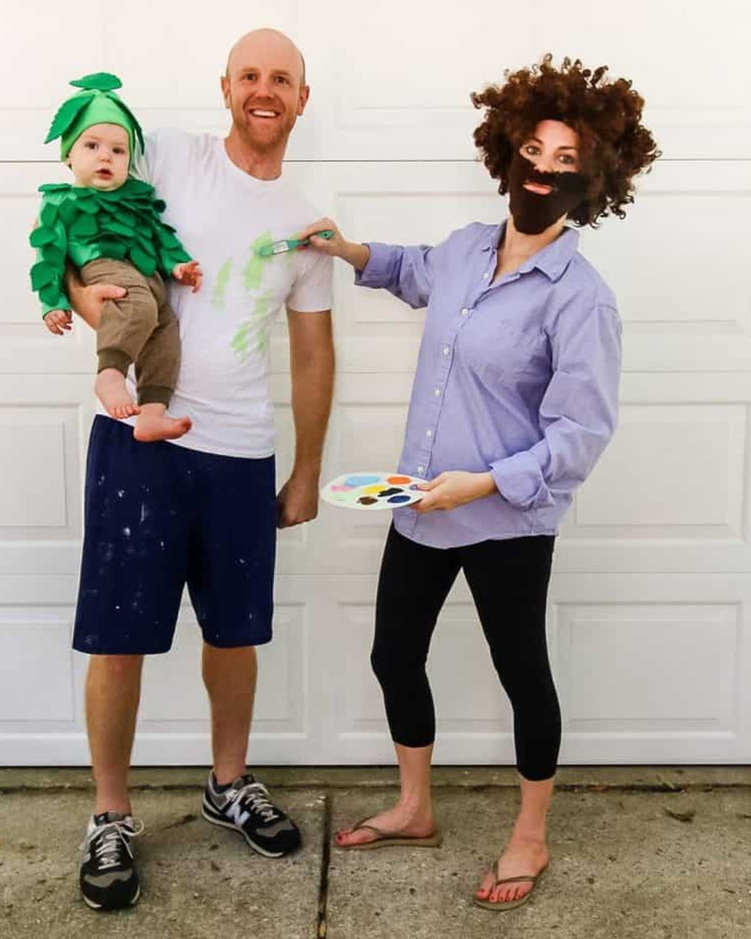 A parent dressed as painter Bob Ross with a curly wig and smock standing next to a toddler wearing a green felt-leaf tree onesie.