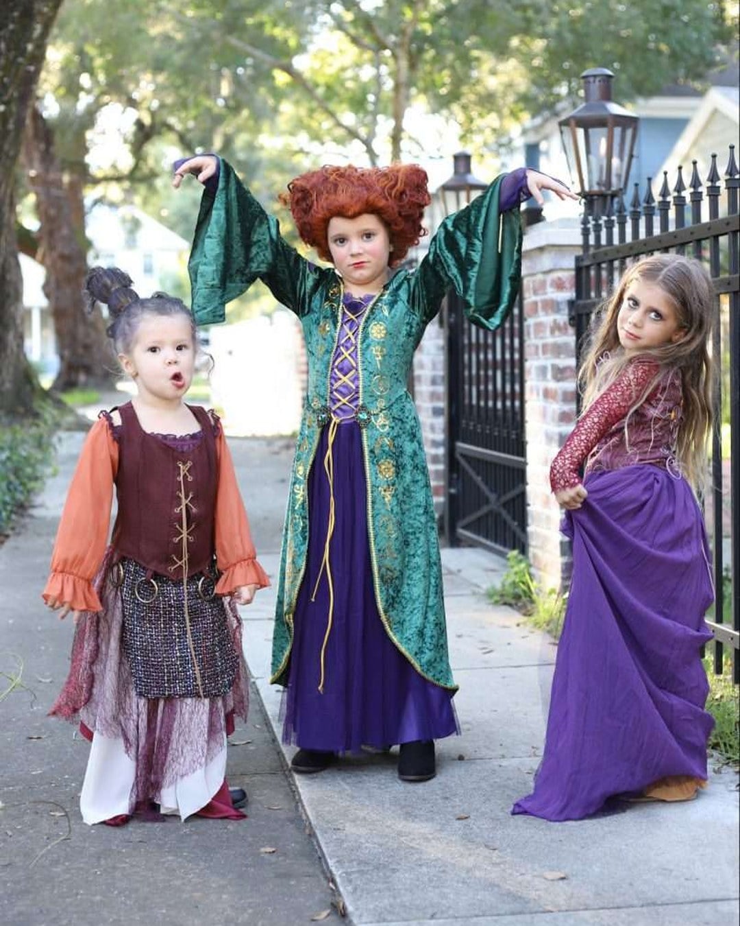 Three young girls dressed as the Sanderson sisters from Hocus Pocus in colorful thrifted dresses, capes, and wild wigs, alongside their matching parents.