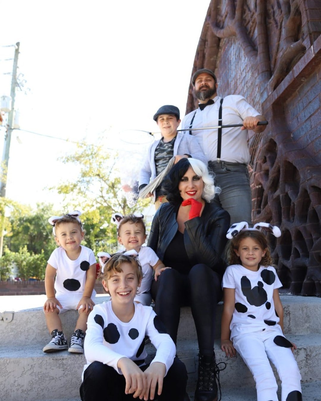 A mom dressed as Cruella de Vil standing with several kids wearing black-and-white spotted dalmatian onesies and ear headbands.