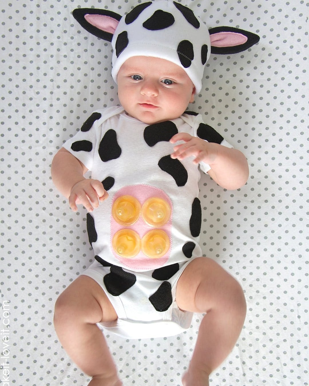 Baby dressed as a spotted cow with a floppy-eared fleece hat and an udder made from baby-bottle nipples on the belly.