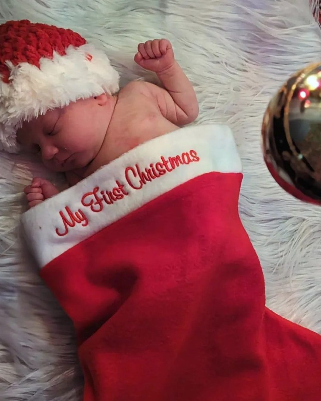A newborn tucked inside an oversized knit Christmas stocking on faux fur under a Christmas tree, face peeking out.