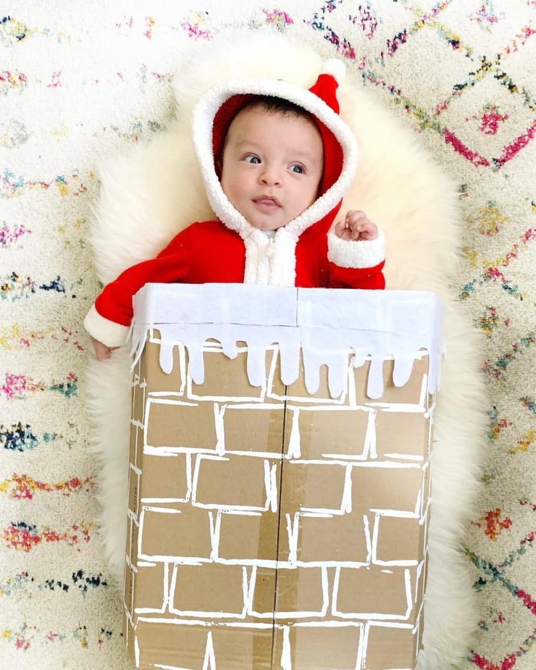 A baby dressed as Santa posed at the base of a DIY cardboard chimney decorated with garland.