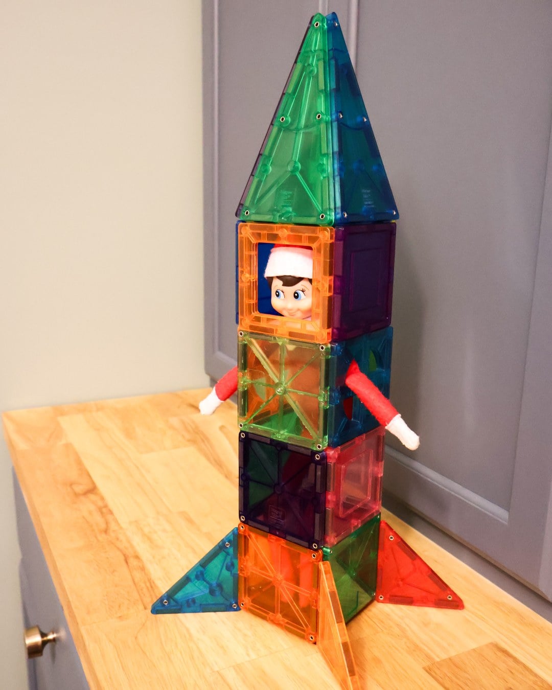 Elf on the Shelf peeking out of a rocket ship built from colorful magnetic building tiles