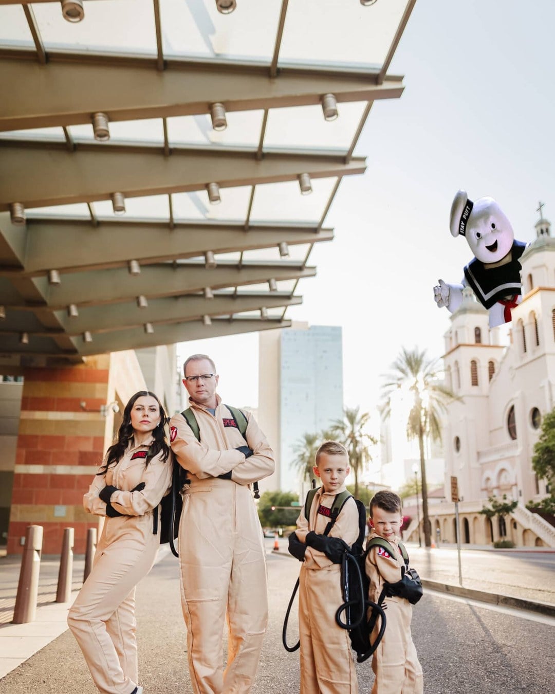 Family dressed as the Ghostbusters team in matching jumpsuits with proton-pack backpacks, including one member as the Stay Puft Marshmallow Man, photographed downtown