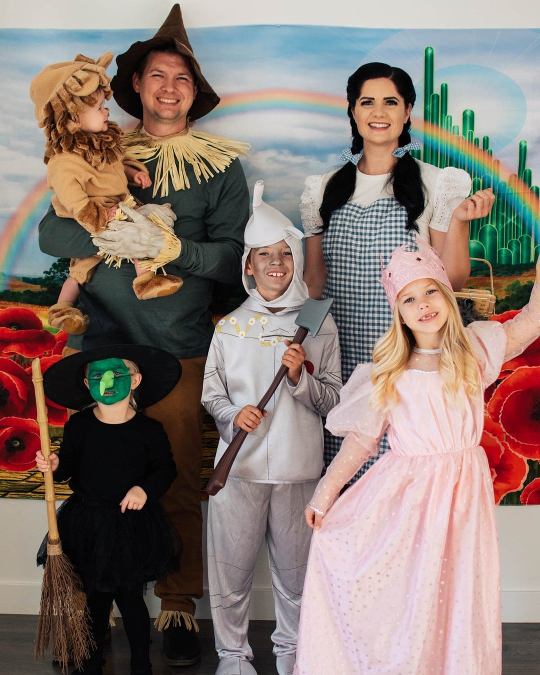 Family of six dressed as Dorothy, the Scarecrow, Tin Man, Cowardly Lion, Glinda, and the Wicked Witch in front of an Emerald City backdrop