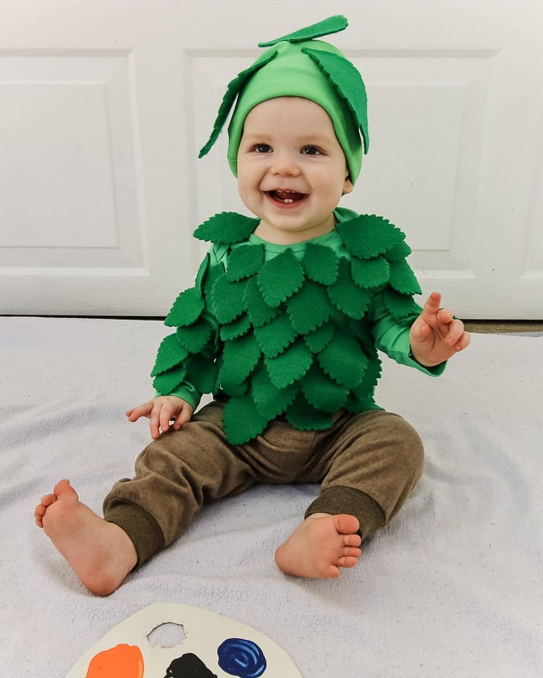 Baby in a green onesie covered in layered felt leaves with a matching leafy green hat and brown pants