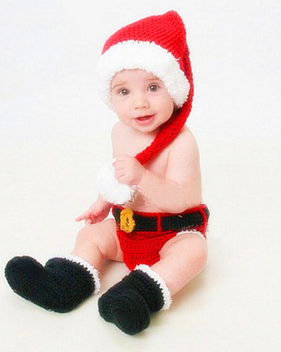 Smiling baby on a white backdrop wearing a red crochet Santa hat, red crochet shorts with belt and black boots