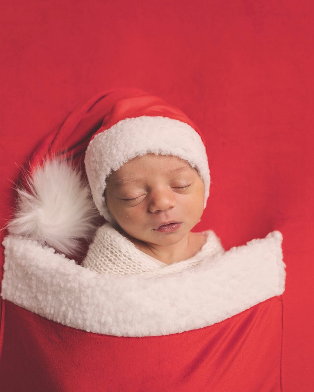 Sleeping newborn in a red Santa hat with fluffy white trim swaddled in cream knit tucked into a red stocking-like pouch