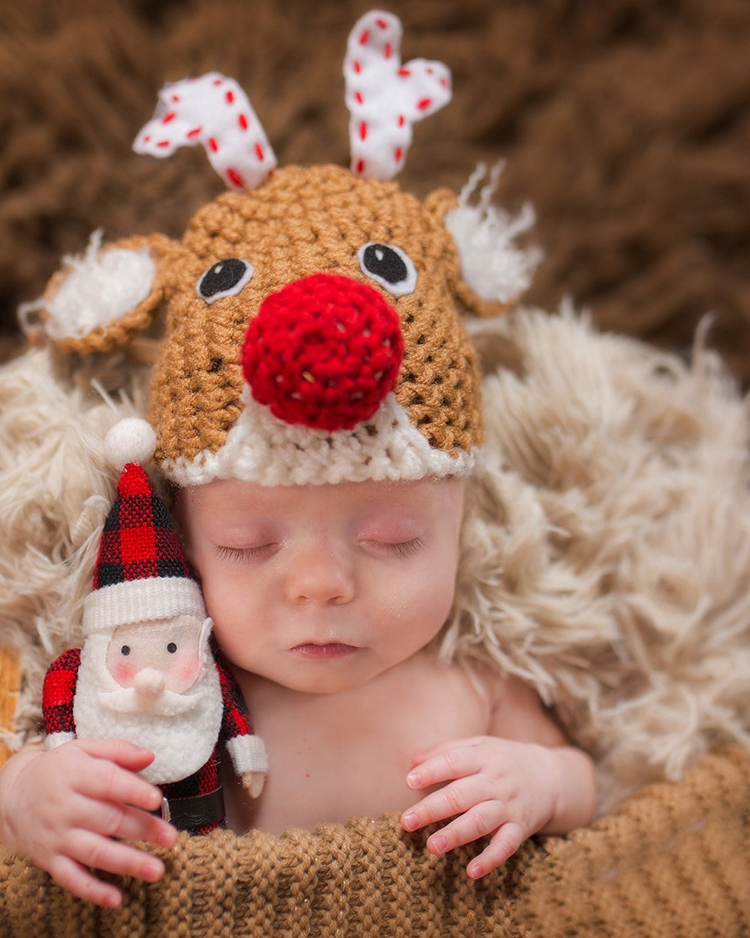 Sleeping baby in a crocheted brown reindeer hat with a big red nose, white antlers and ears in a fluffy basket
