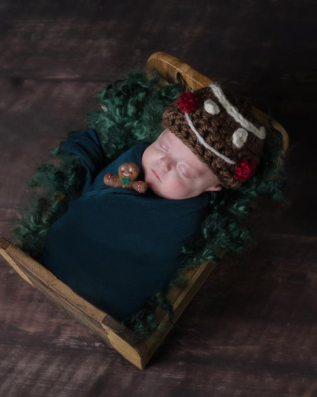 Sleeping newborn in a knitted brown gingerbread-man hat with white icing trim and red buttons, swaddled in a crib