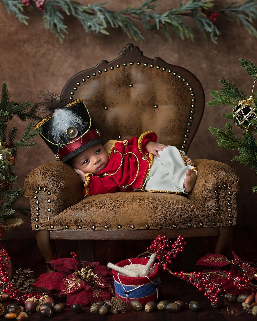 Newborn in a red jacket with gold trim, white trousers and a tall black soldier hat on a vintage armchair