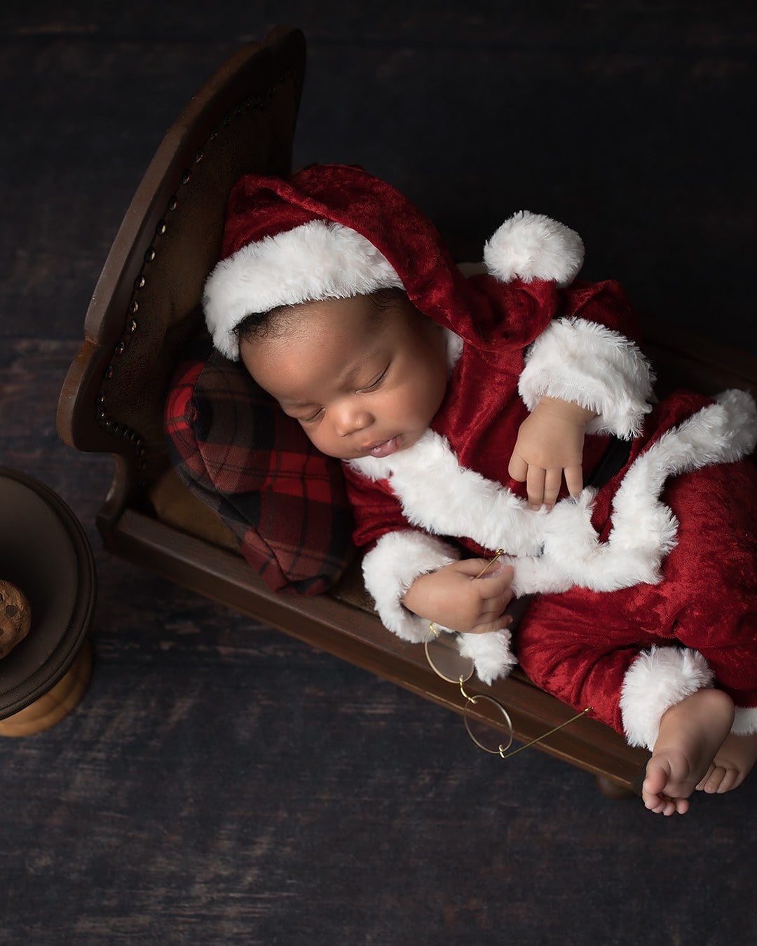 Sleeping newborn in a red velvet Santa outfit lying in a small wooden sleigh-style bed against a dark backdrop