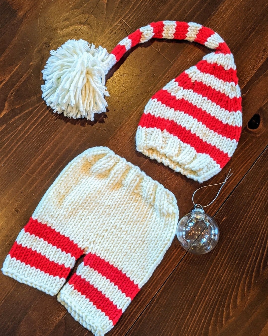 Flat-lay of a red and cream striped knitted elf hat with a pom-pom and matching striped shorts beside a glass ornament