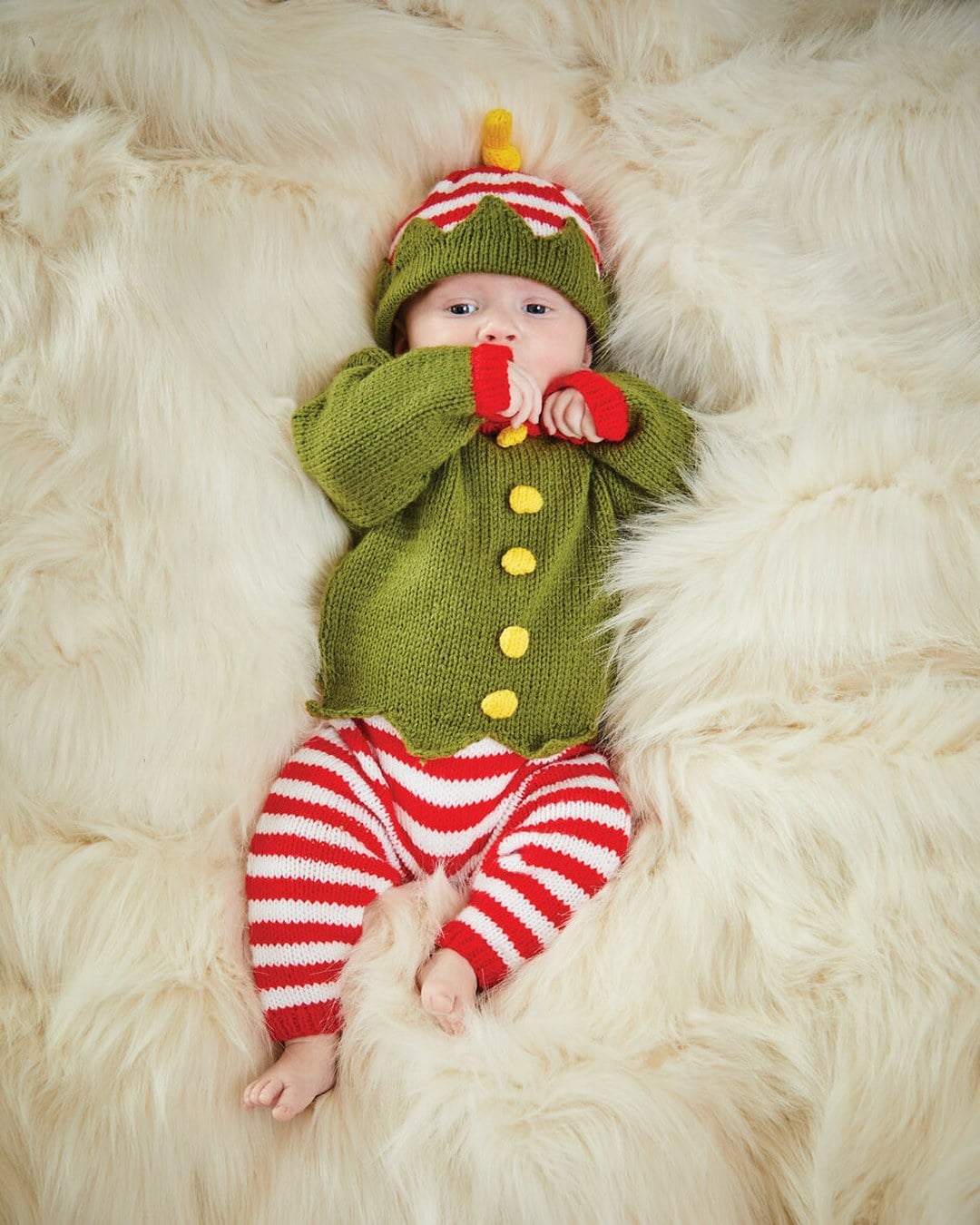 Young baby on a cream faux-fur rug wearing a hand-knitted green elf sweater, striped trousers and a knit elf hat