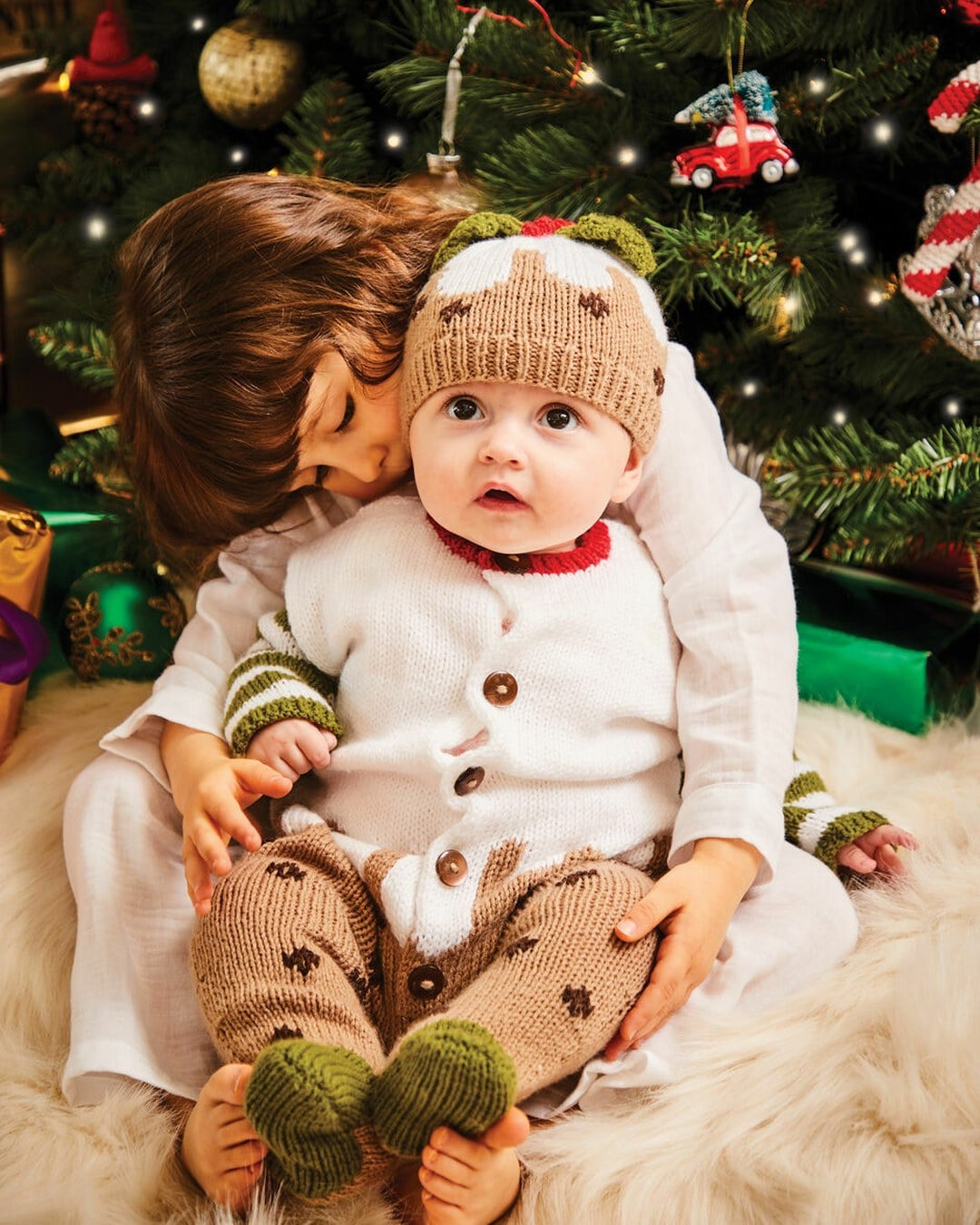 Baby in a knitted Christmas-pudding hat, white cardigan and brown trousers sitting on a fur rug by a tree