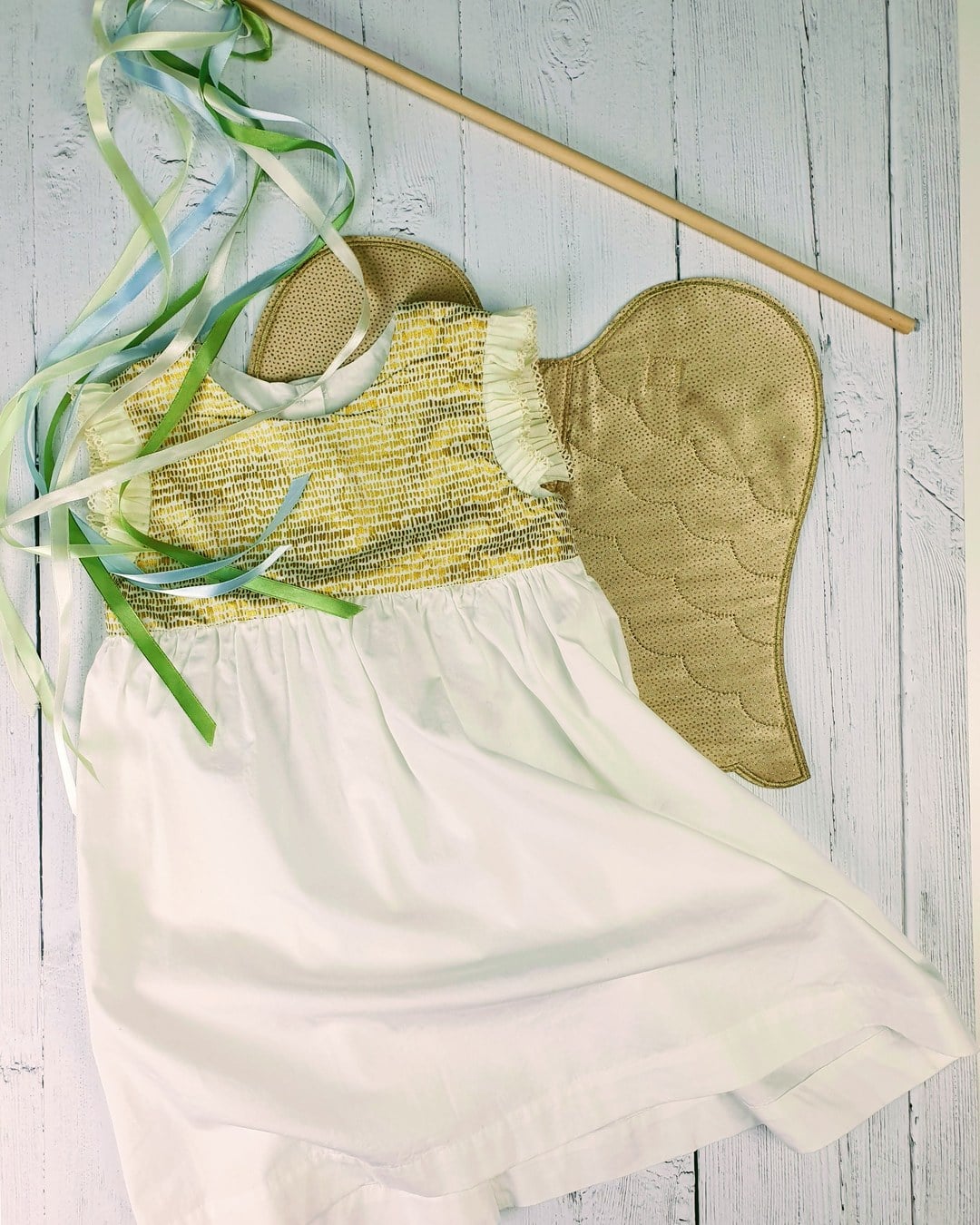Flat-lay of a toddler dress with a gold smocked bodice and white skirt next to a gold quilted wing and ribbon wand