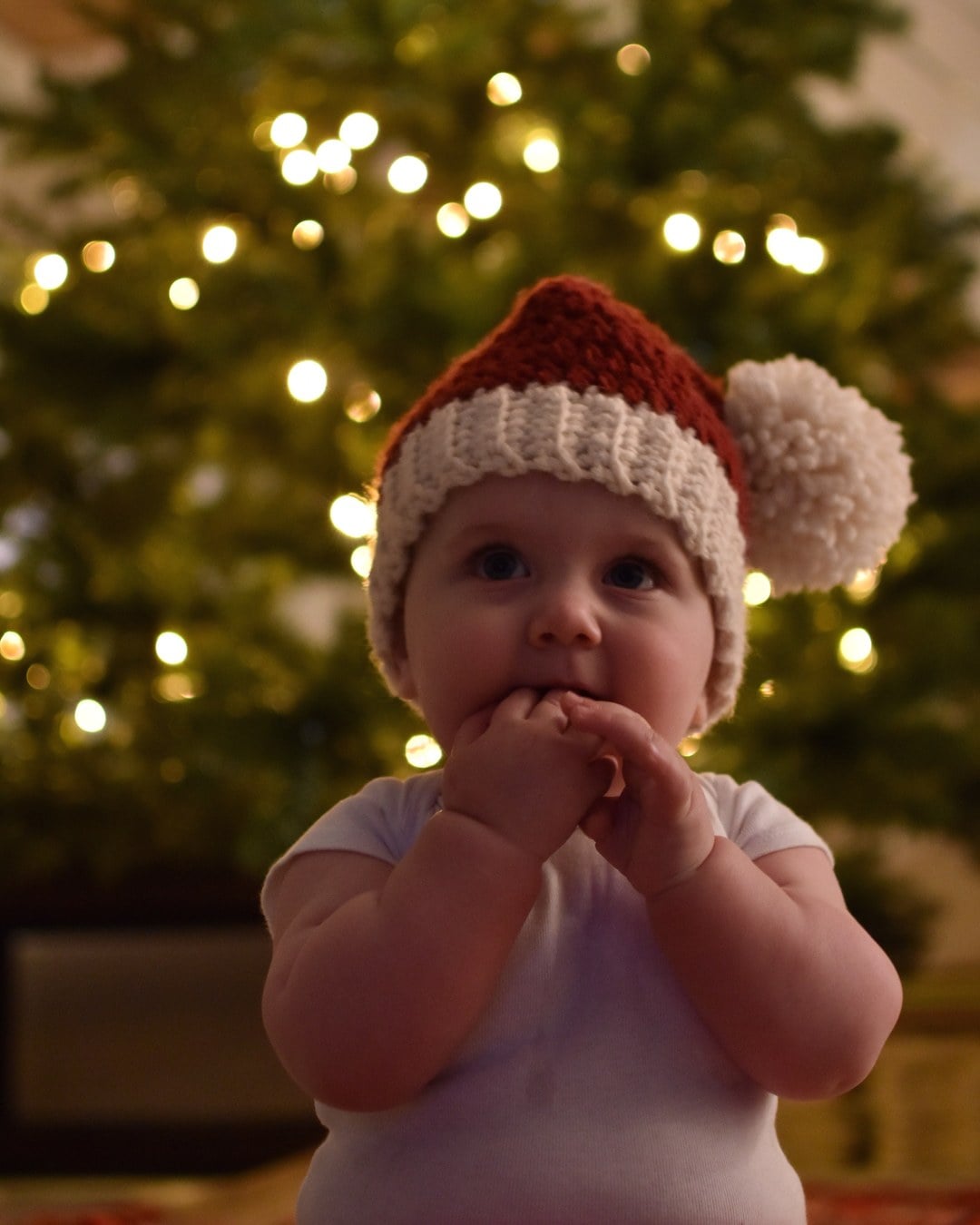 Baby in a crocheted red Santa hat with a big pom-pom and a white onesie in front of a lit Christmas tree