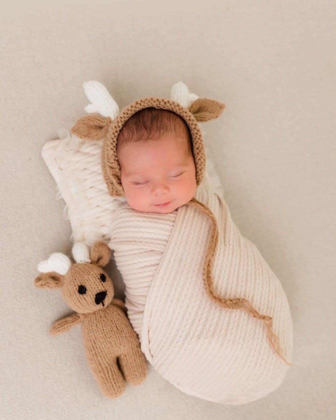 Sleeping newborn swaddled in cream wearing a brown knit bonnet with ears and small white antlers beside a knitted reindeer toy