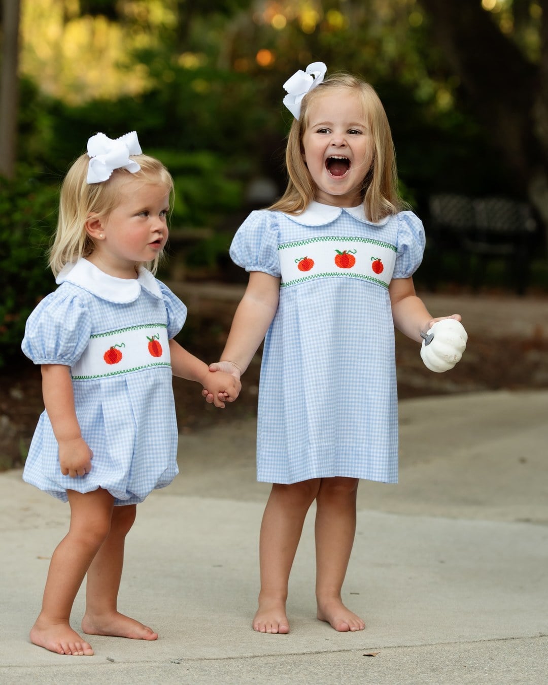 Two toddler sisters in matching gingham smocked outfits with pumpkin embroidery holding hands outdoors
