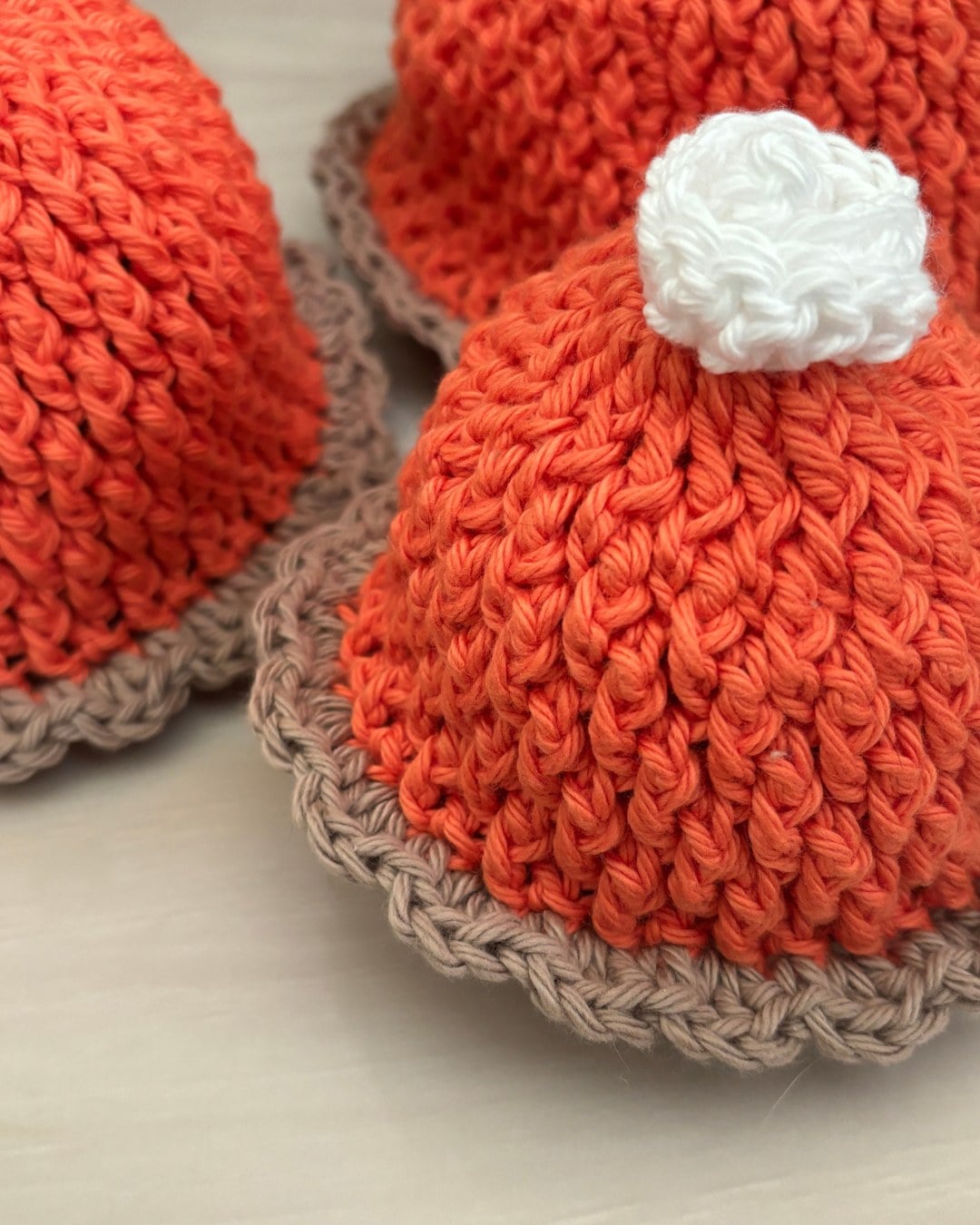 Three orange crochet hats with tan scalloped brims and white pom-poms styled like pumpkin pies