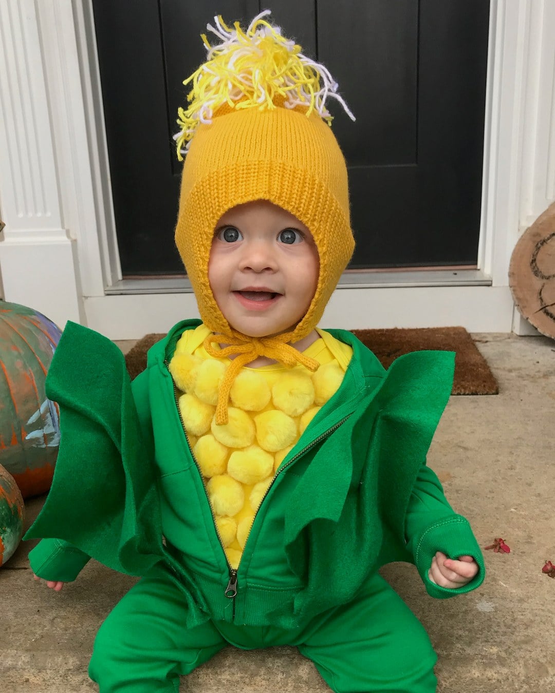 Baby in a corn costume with a yellow knit hat, yellow pom-poms on the chest and green felt husks
