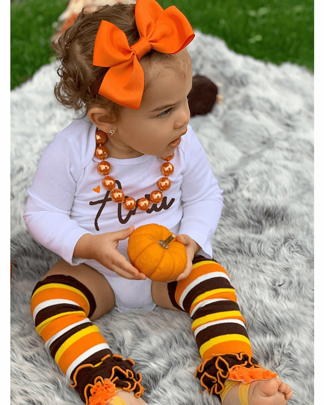 Brown, orange and yellow striped ruffle leg warmers on a toddler wearing a white bodysuit and orange bow