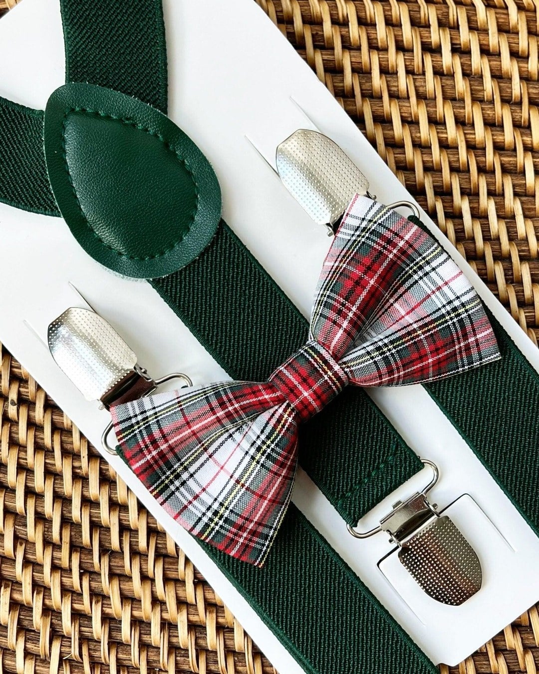 Dark green suspenders with a red, white and green plaid bow tie on a white card over a woven surface