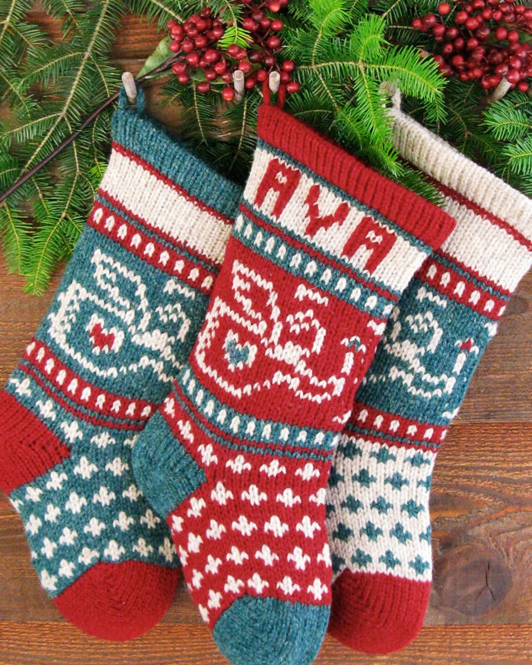 Three Fair Isle-style knit stockings, one with the name AVA and an angel motif, hanging against greenery
