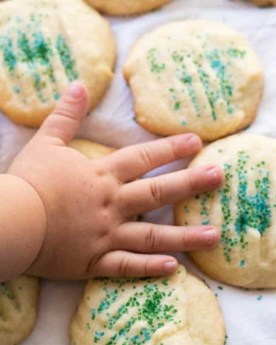 A baby's hand resting on baked sugar cookies topped with green and blue sprinkles