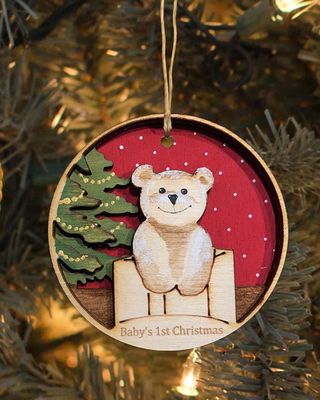 A layered hand-painted wooden ornament with a teddy bear and a tree, reading Baby's 1st Christmas, hanging on a lit tree