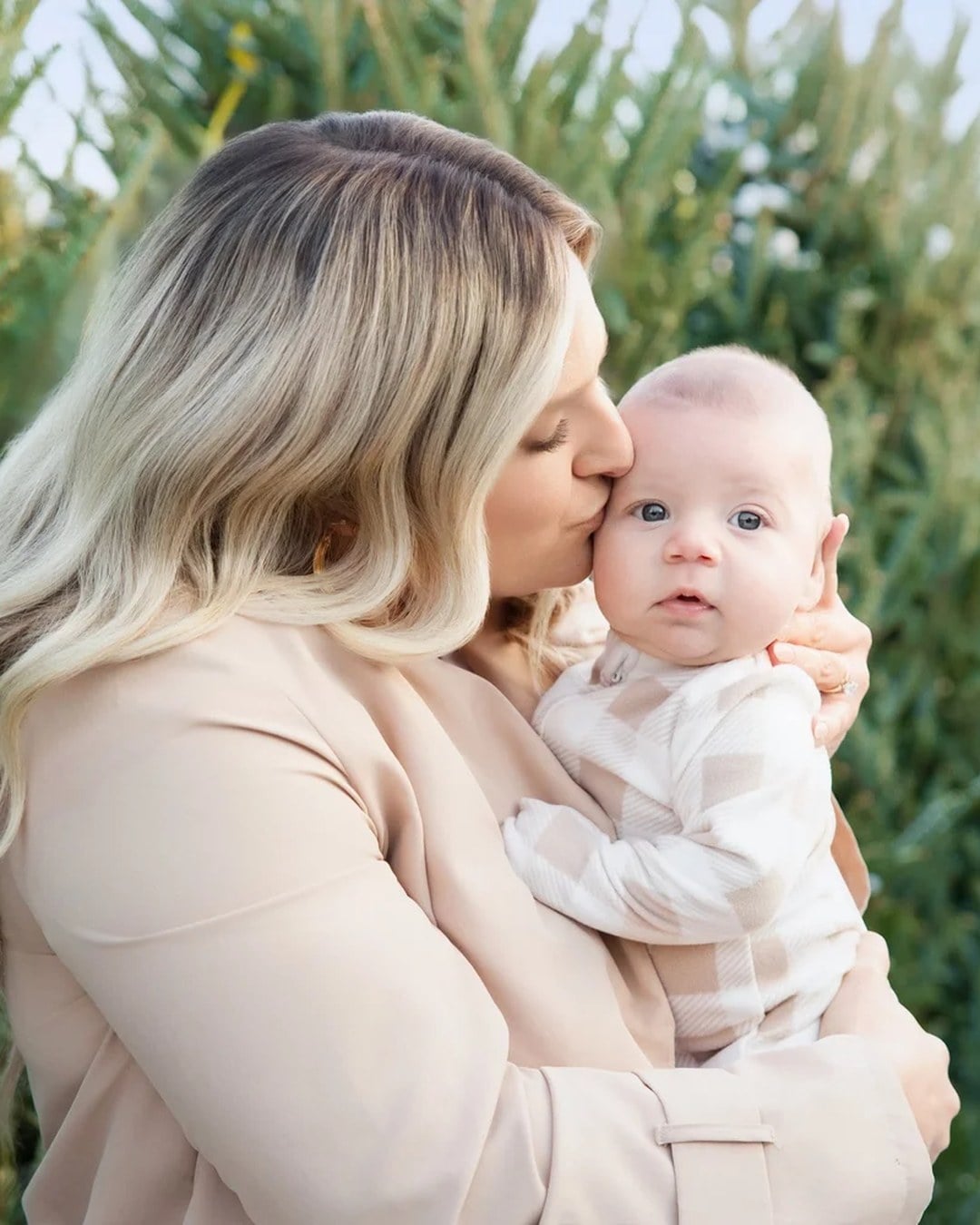 A woman kissing the cheek of a wide-eyed baby with Christmas trees behind them at a tree farm