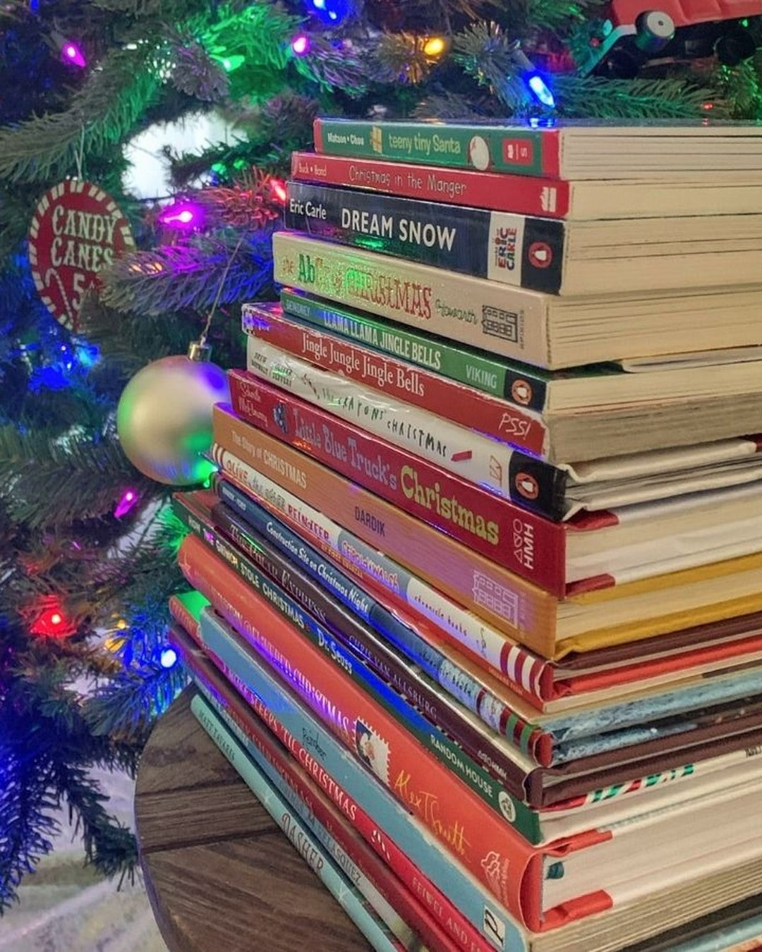 A stack of wrapped holiday books beside children reading in front of a Christmas tree