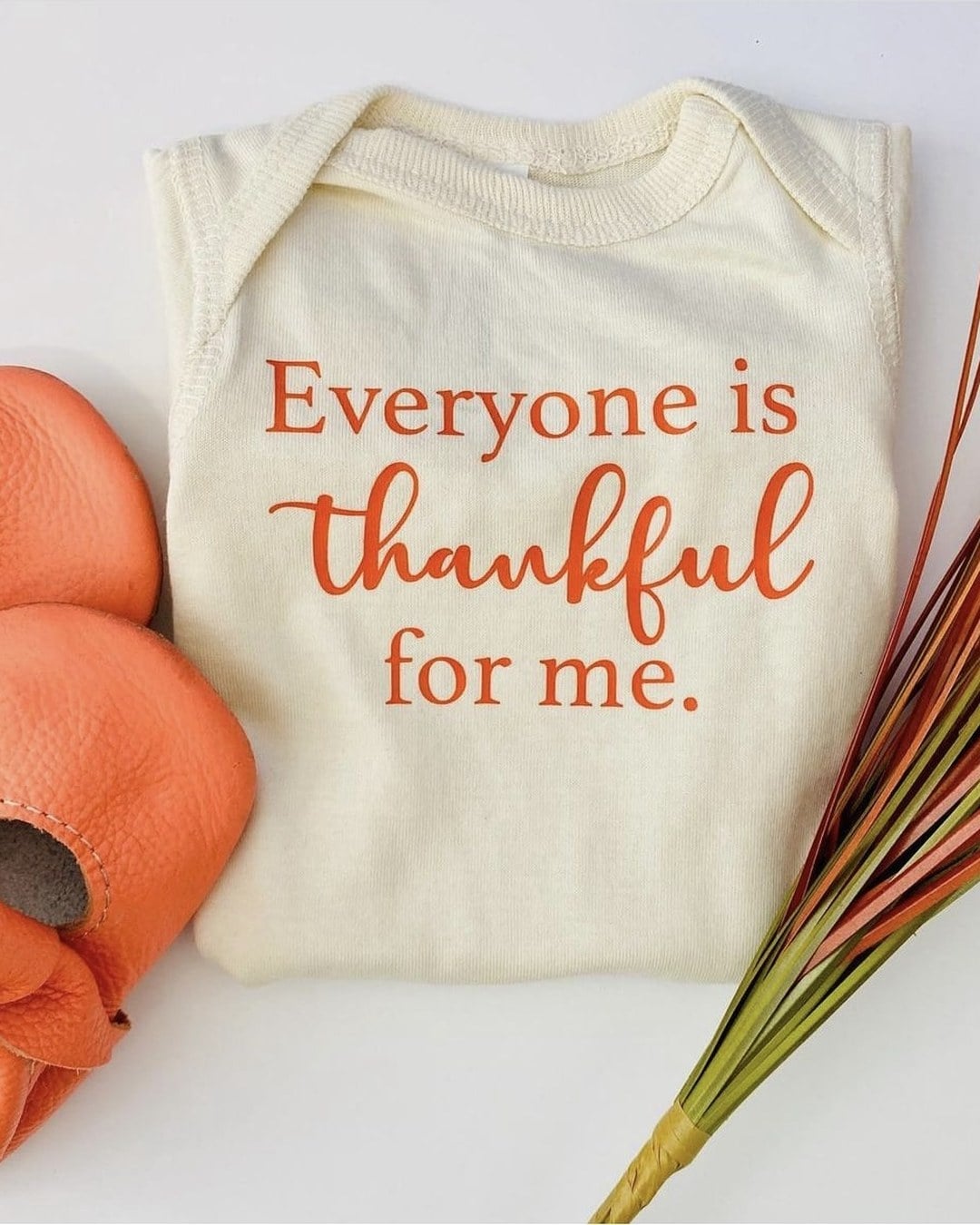 A flat-lay of a cream onesie printed 'Everyone is thankful for me.' beside orange baby booties and a bundle of orange and green fall grass