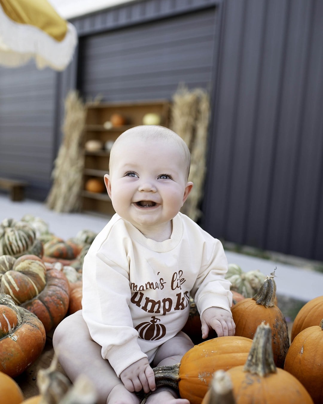 A smiling baby in a cream pumpkin-print sweatshirt romper sitting among orange and striped pumpkins in front of a gray barn with hay bales