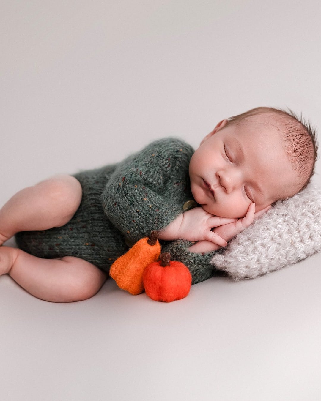 A sleeping newborn in a green knit romper lying on its side on a light backdrop, resting on a small knit pillow beside two felt pumpkins